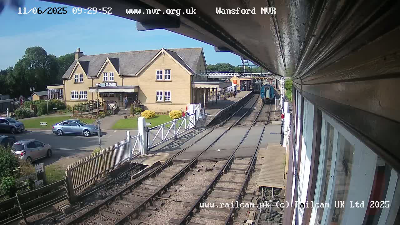 A train departs a station on a sunny day, passing a two-story building and several cars parked nearby.