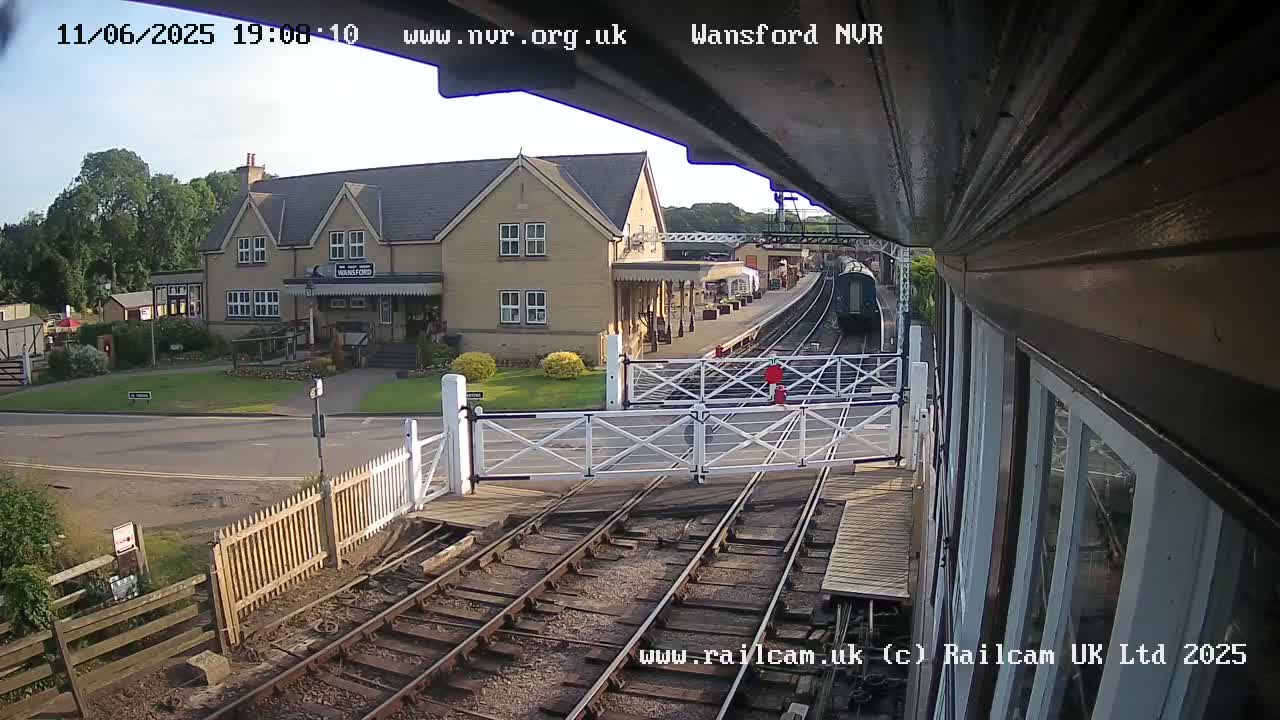 A train approaches a station building on a sunny afternoon, passing through a level crossing.