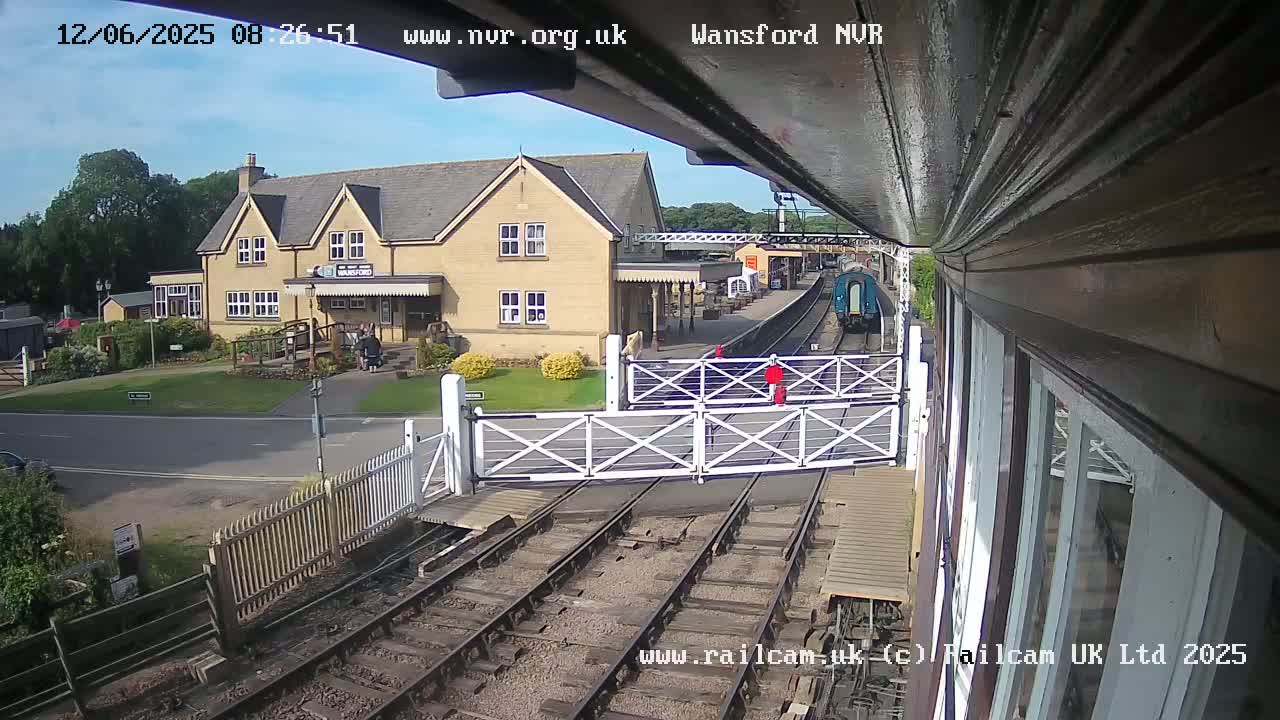 A train approaches a railway station on a sunny day, passing through a level crossing gate near a light-colored building.