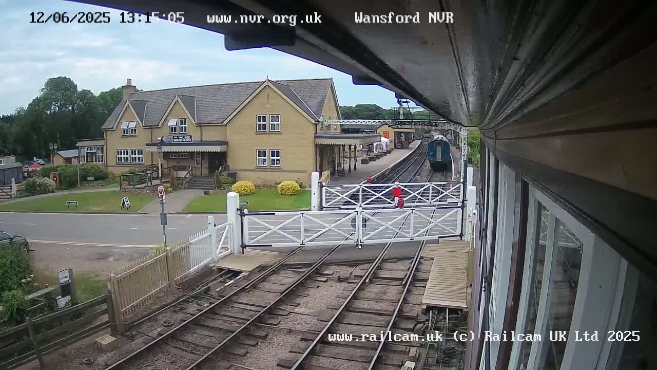 A train approaches a railway station on a partly cloudy day, passing through a level crossing with gates down.
