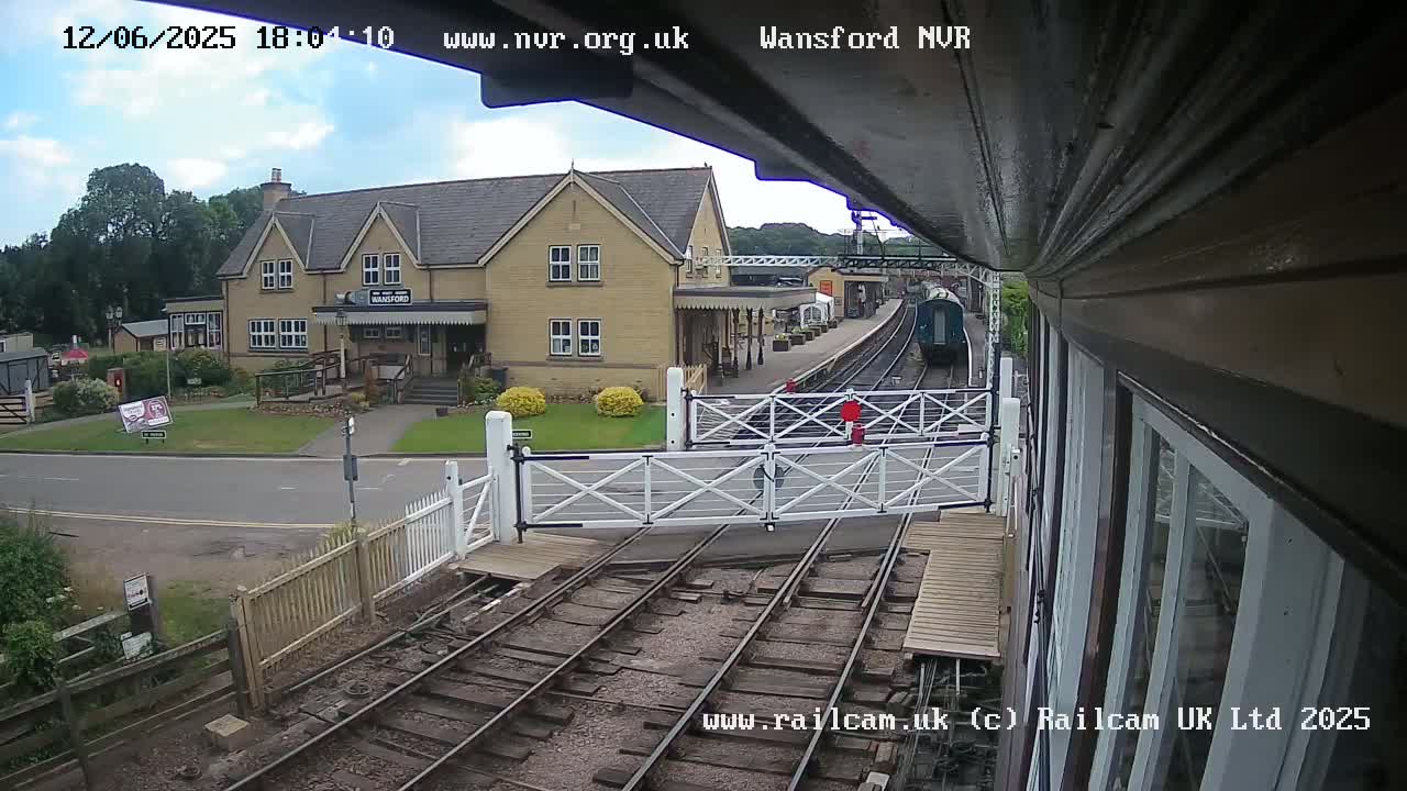 A train is arriving at a railway station on a partly cloudy day, passing through a level crossing with white gates.