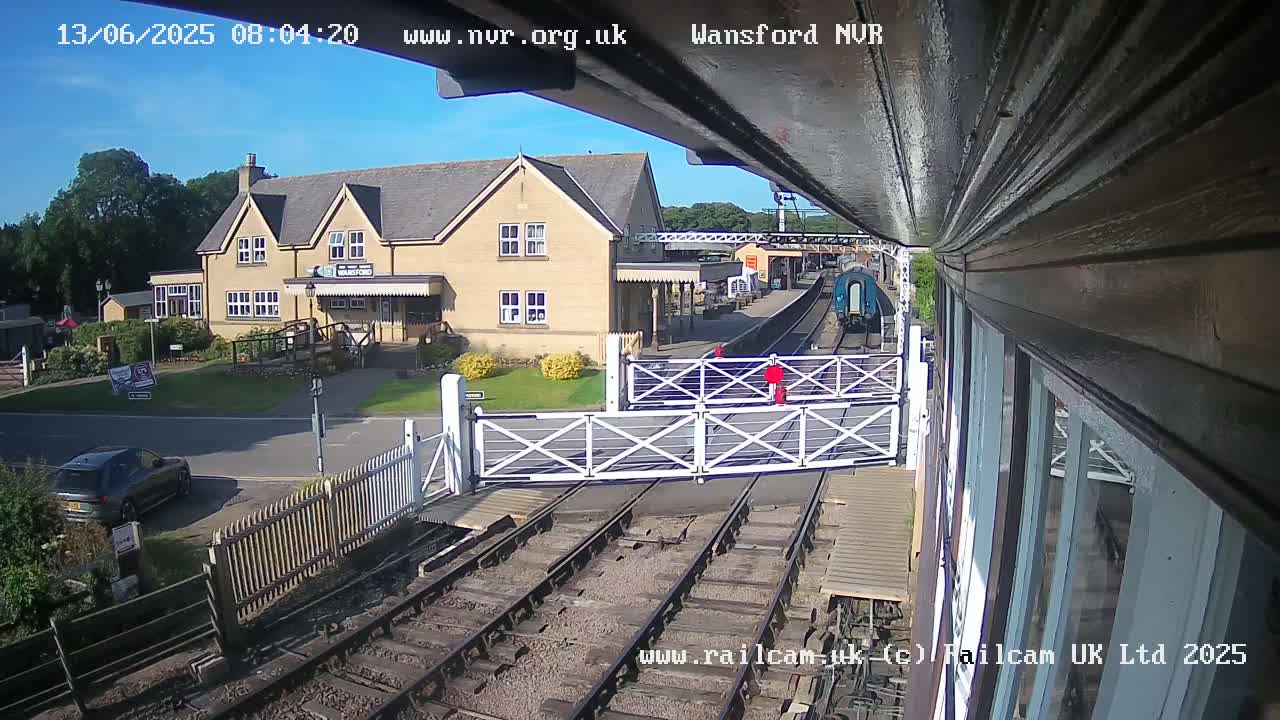 A train is arriving at a station on a sunny day, passing through a level crossing near a two-story building.