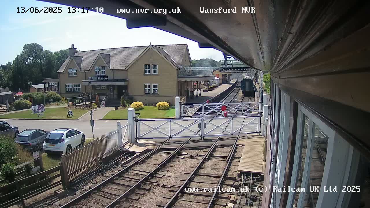 A train approaches a station on a sunny day, passing through a level crossing near a building with a grassy area and parked cars.