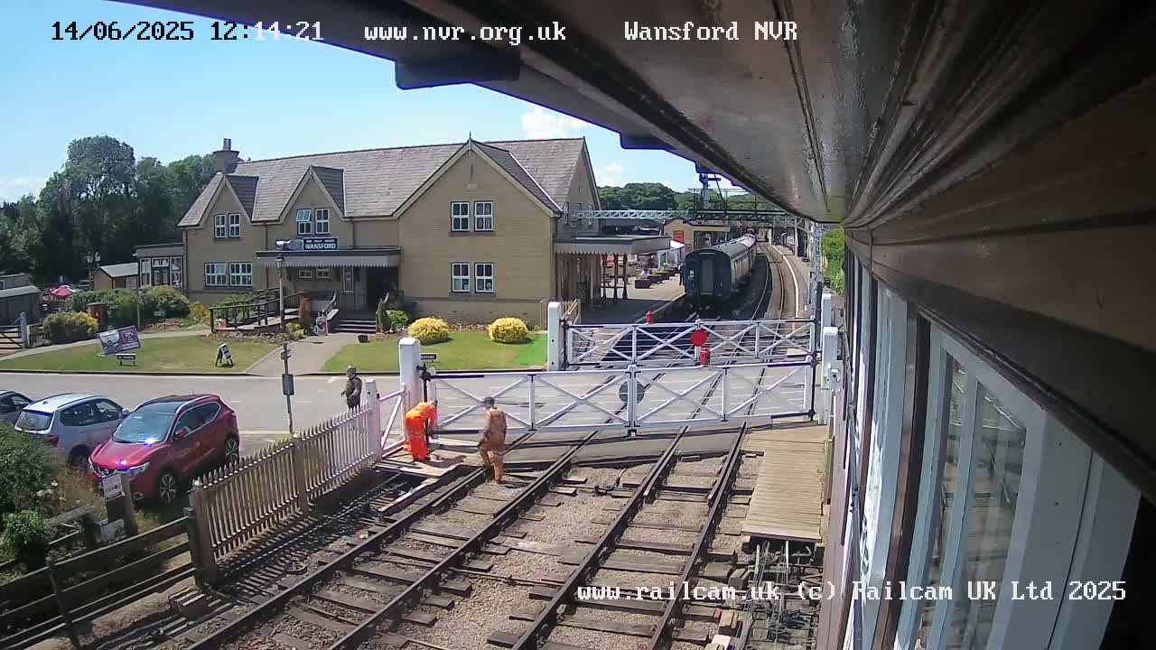 A train is arriving at a station on a sunny day, with two people in orange vests working near the tracks and several cars parked nearby.