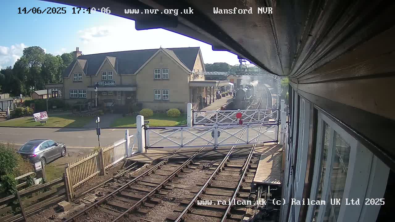 A steam train is departing from a station on a sunny day, passing through a level crossing near a building and a car.