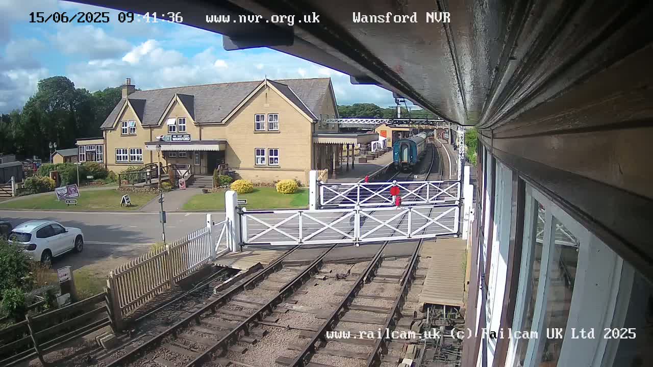 A train is arriving at a station on a partly cloudy day, with a building and a car visible in the foreground.