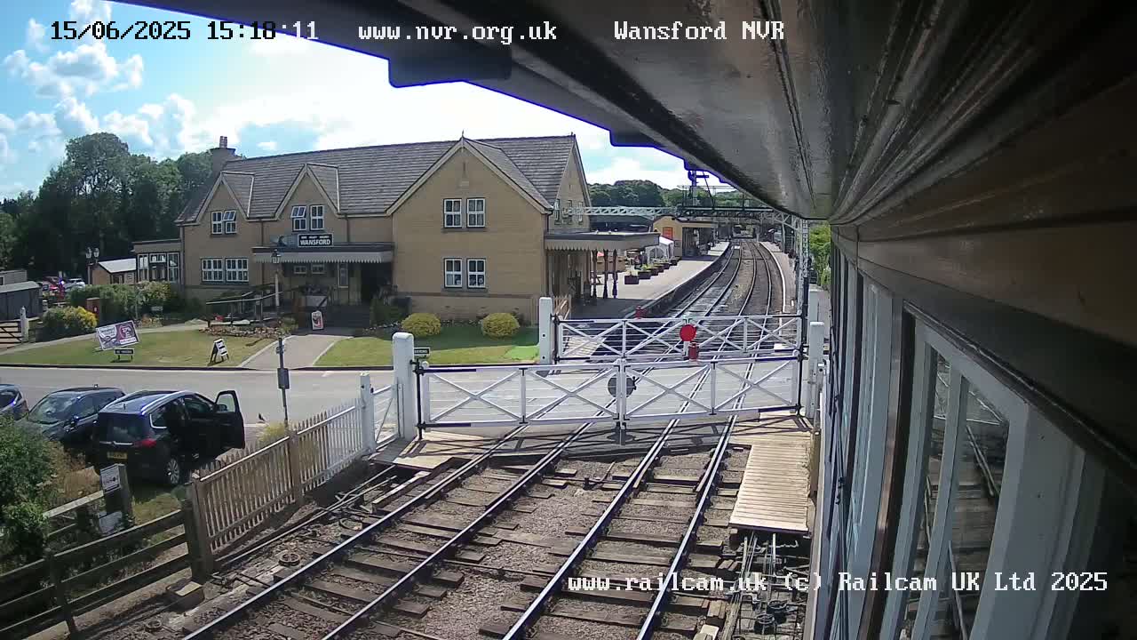 A train station viewed from a train, under sunny skies, shows a two-story building and railway tracks with crossing gates.