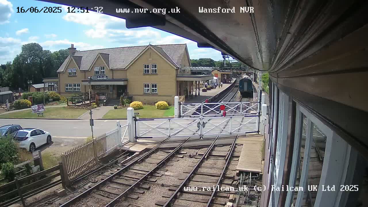 A train approaches a station on a sunny day, passing through a level crossing in front of a two-story building.
