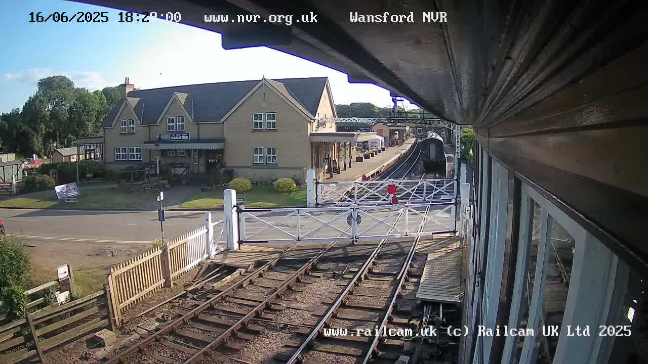 A train approaches a railway crossing in front of a building under a mostly sunny sky.