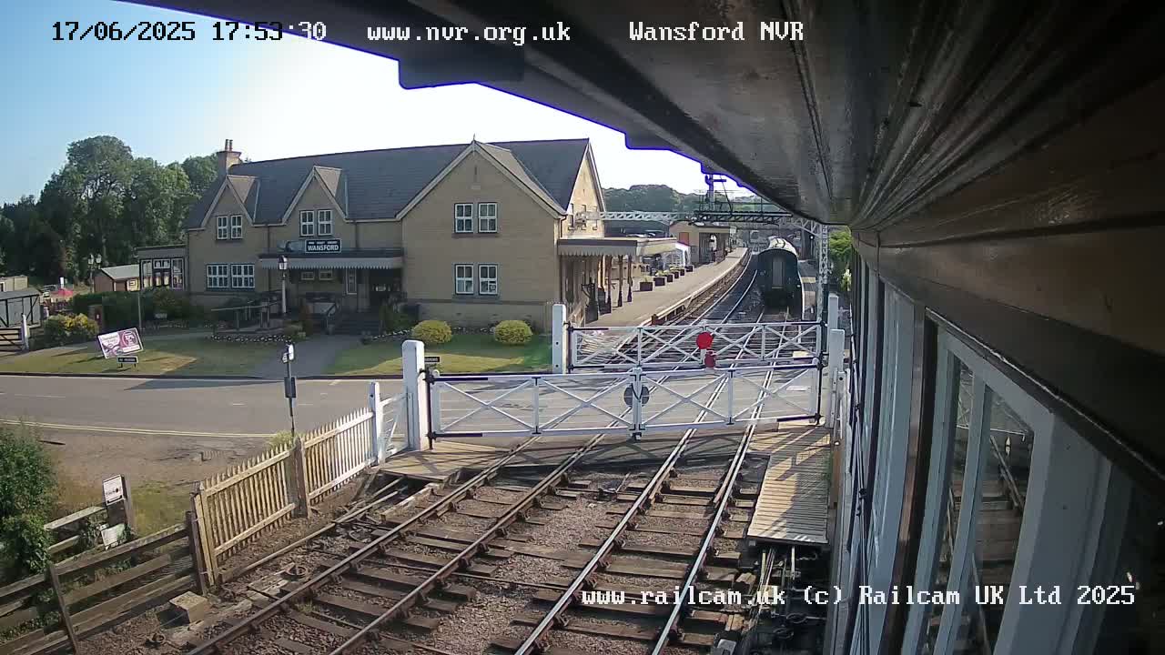 A train station with a two-story building, a train on the tracks, and level crossing gates in sunny weather.