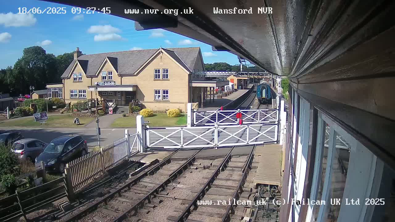 A sunny day at a train station shows a train arriving at a platform next to a light-colored building with a parking lot.