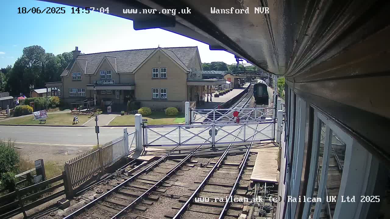 A sunny day at a train station shows a train approaching the platform, passing through a level crossing, with a building in the background.