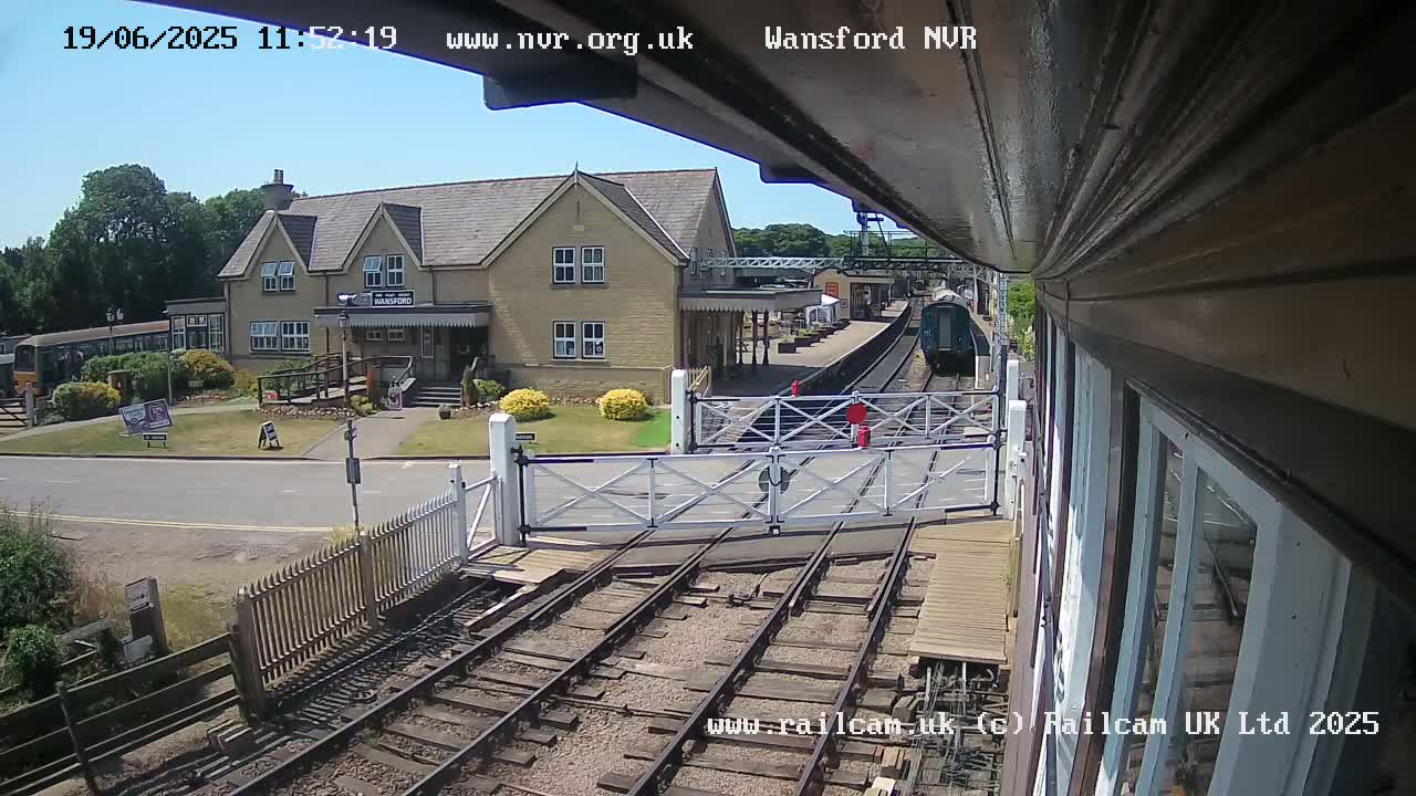 A train pulls away from a station on a sunny day, passing through a level crossing in front of a light-colored building.
