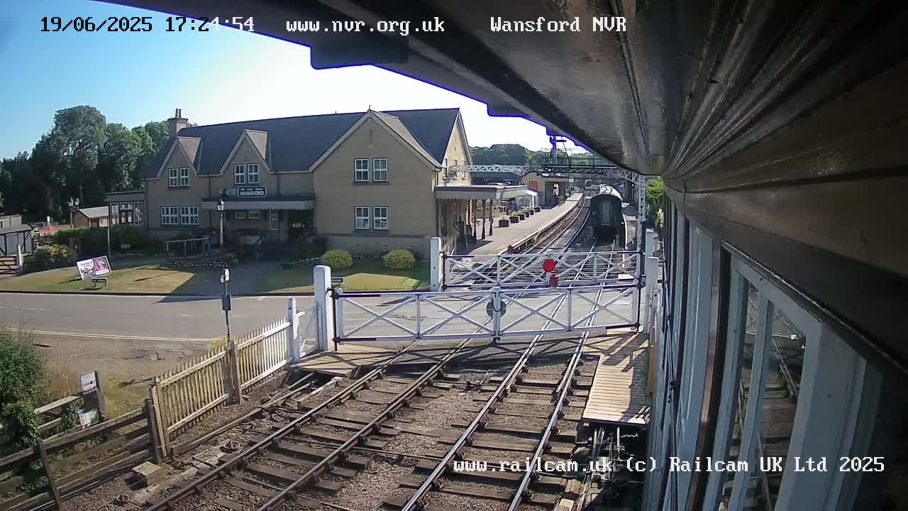 A sunny view from a train shows a railway station building, train tracks, and a crossing gate with a train in the distance.
