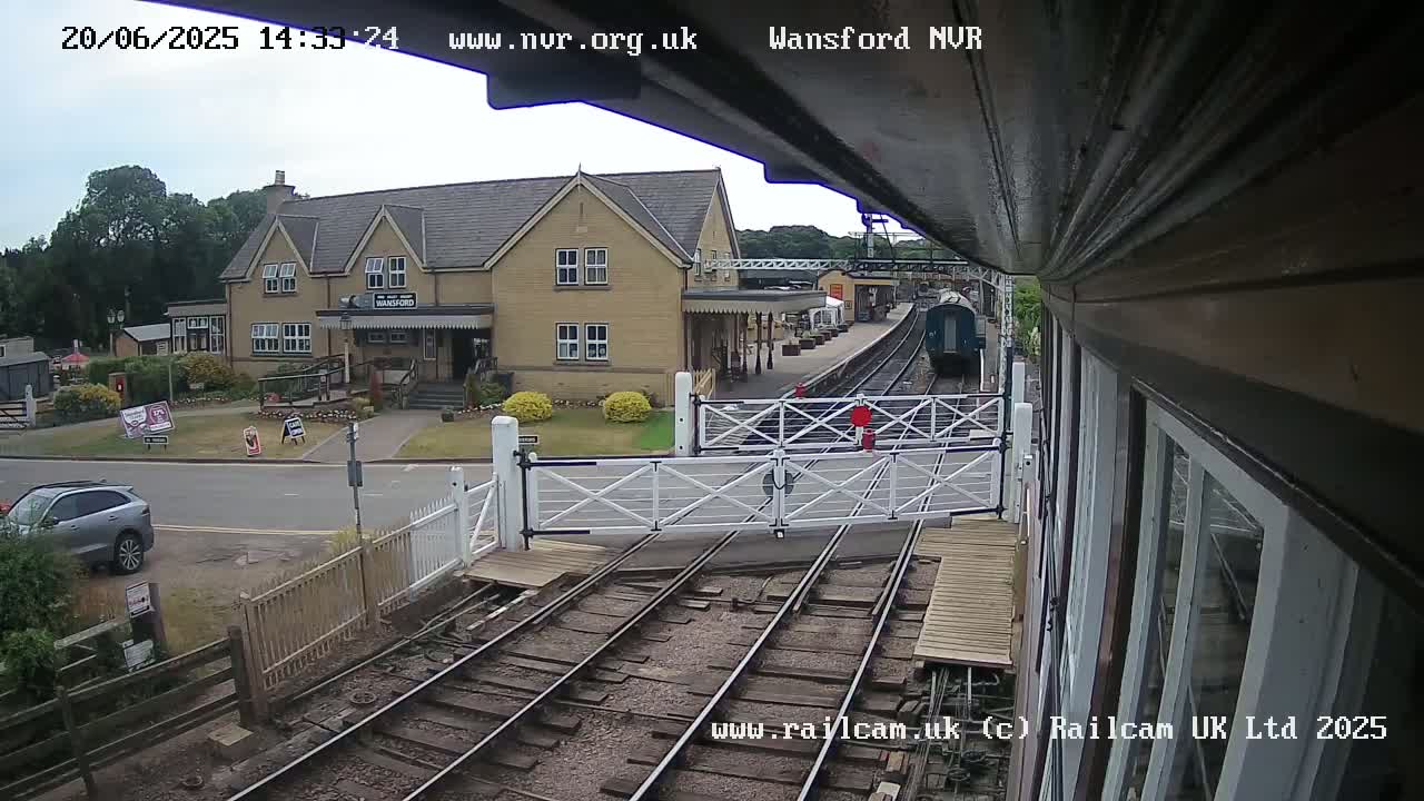 A train is approaching a station on a partly cloudy day, with a building and a few cars visible.
