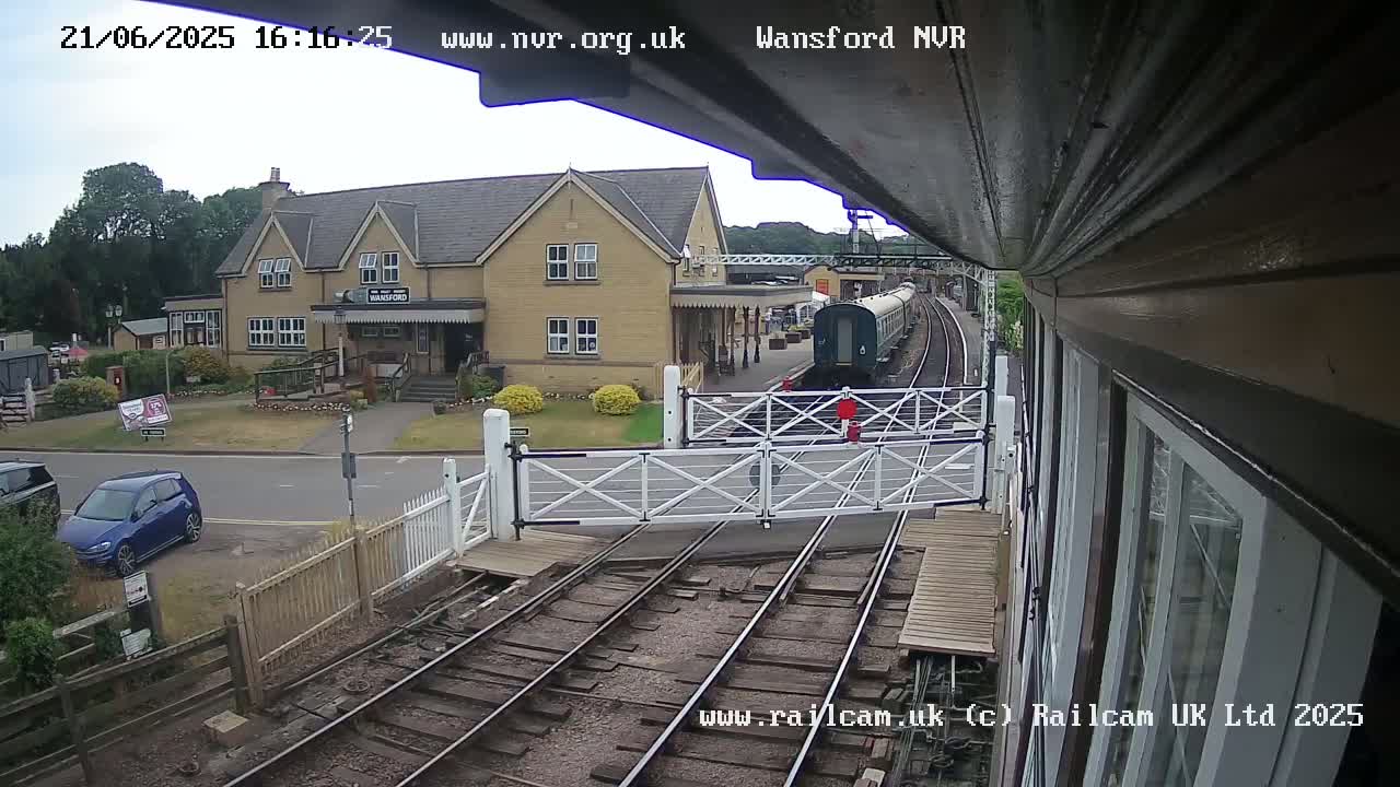 A train approaches a station building on an overcast day, passing through a level crossing with a car parked nearby.