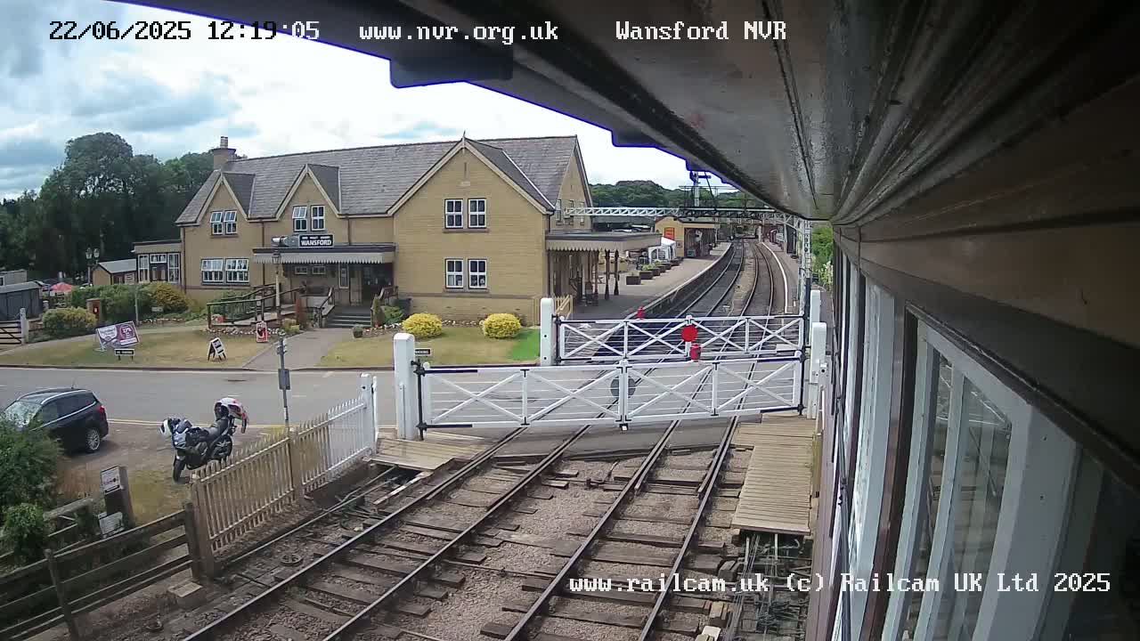 A partly cloudy day shows a train station with a tan building, railroad tracks, and a level crossing gate; two motorcycles are parked nearby.