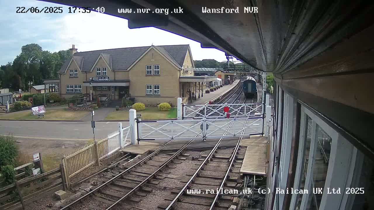 A train is departing from a station on a partly cloudy day, passing through a level crossing with a building visible in the background.