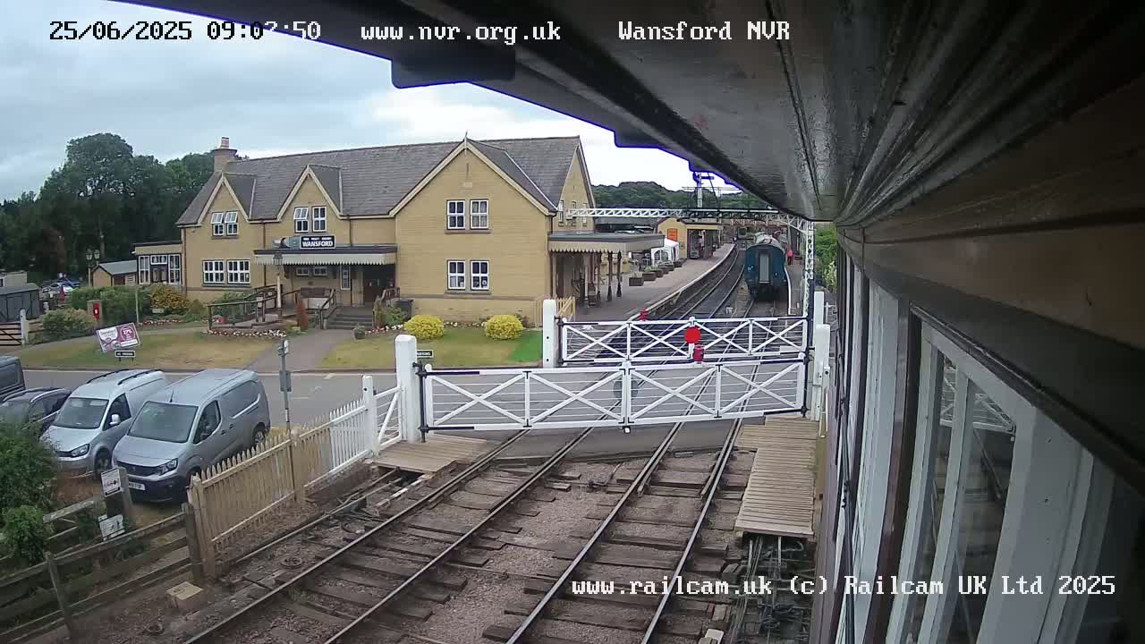 A train is arriving at a station with a two-story building and several parked vans, under a mostly cloudy sky.