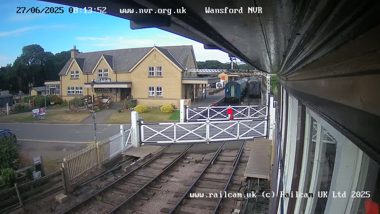 A train is stopped at a station with a light-colored building and closed crossing gates under a partly cloudy sky.