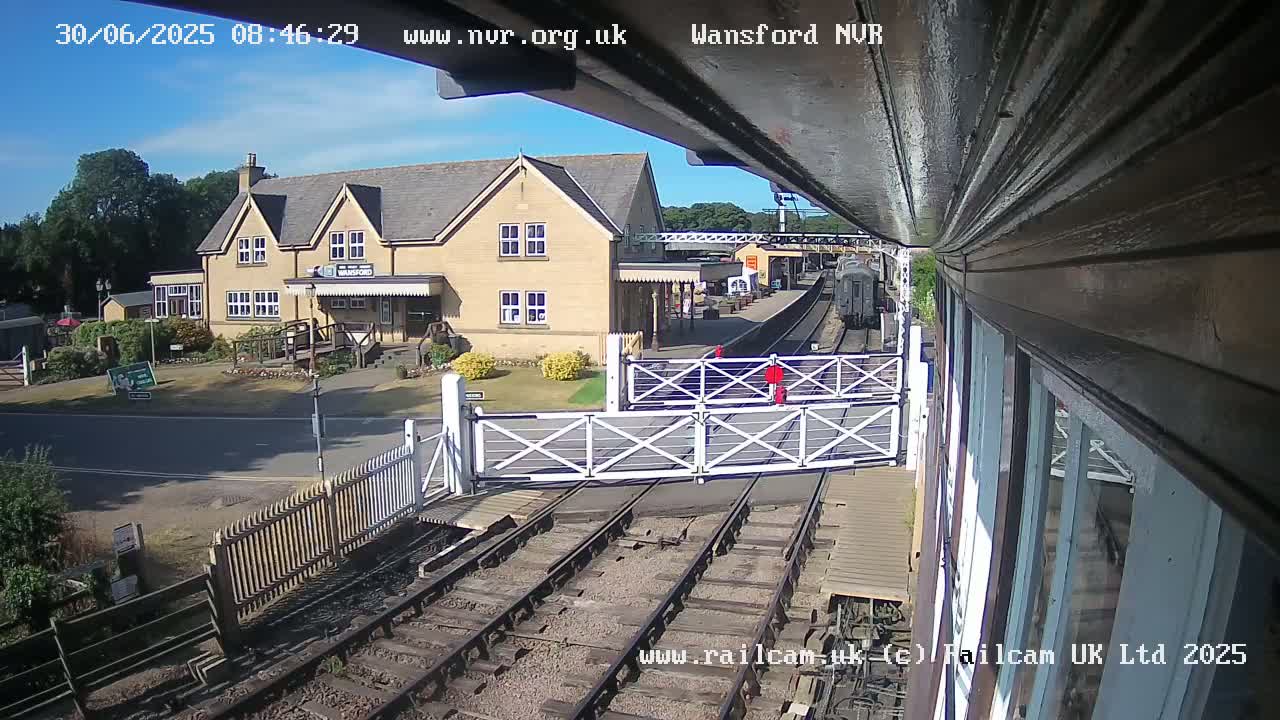 A train approaches a station on a sunny day, passing through a level crossing gate in front of a light-colored stone building.