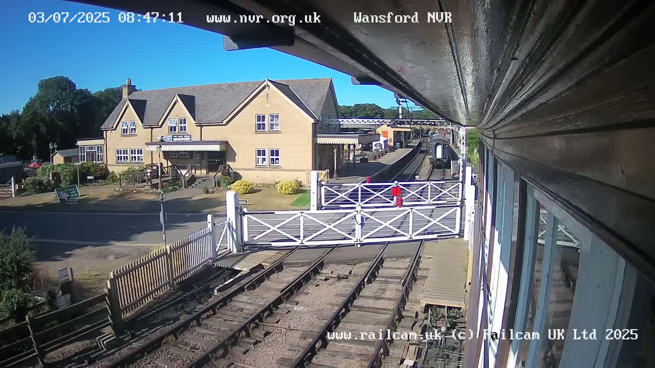 A train approaches a railway station on a sunny day, passing through a level crossing gate near a stone building.