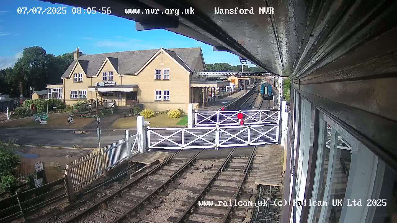 A train departs a railway station on a sunny day, passing through a level crossing with gates down.