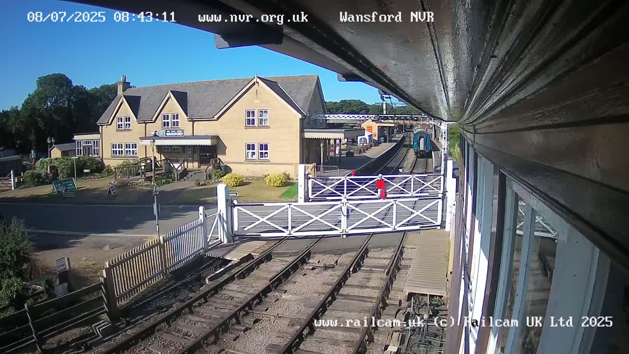 A train approaches a railway station on a sunny day, passing through level crossing gates.