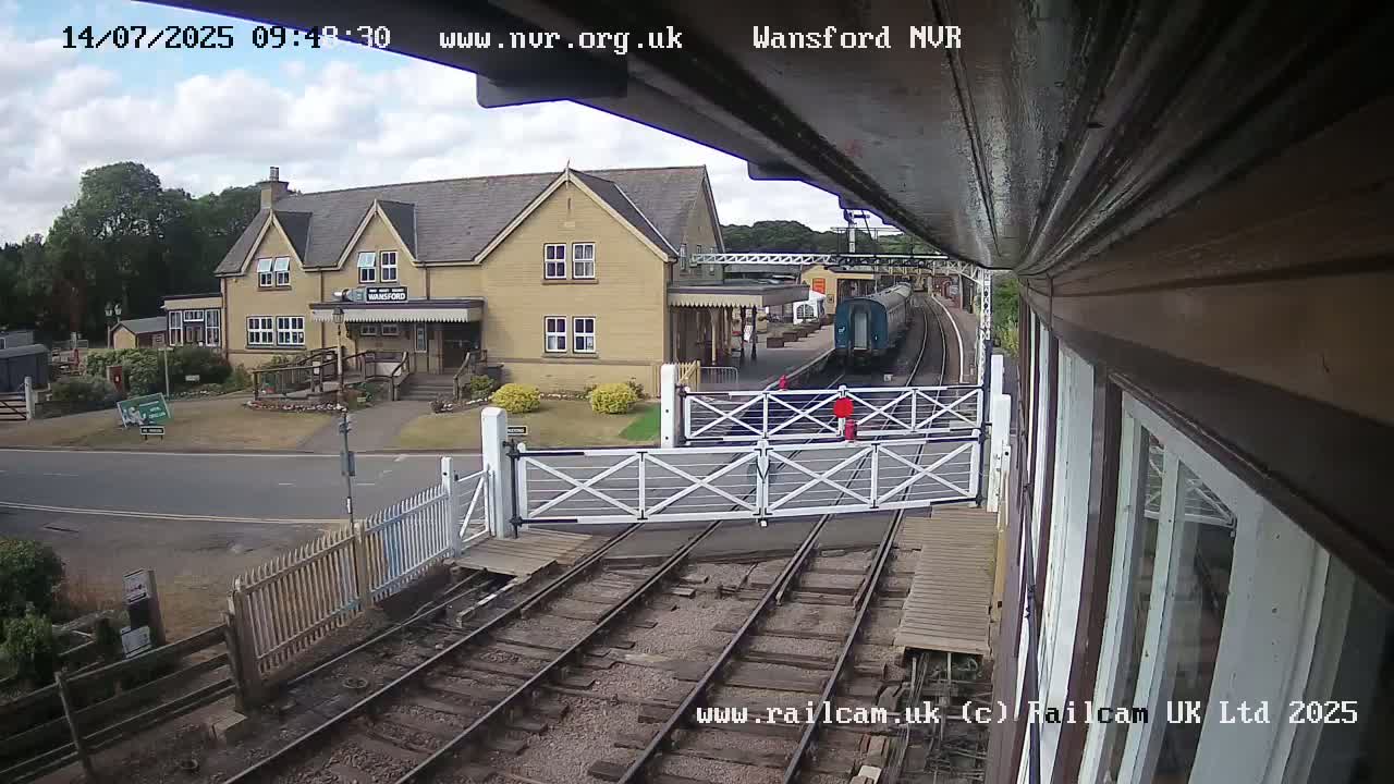 A train passes a station building on a partly cloudy day, with level crossing gates down.