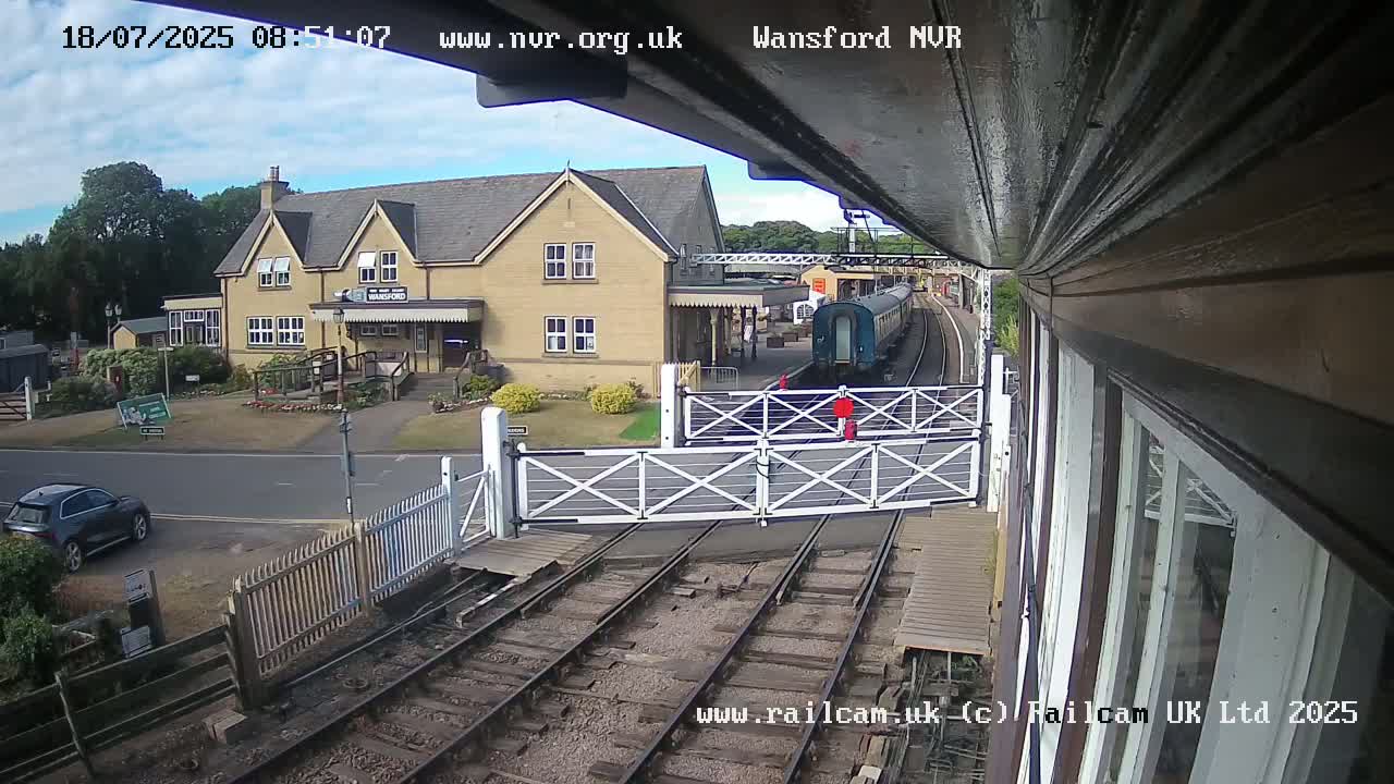 A train departs from a station building on a partly sunny day, passing through a level crossing.