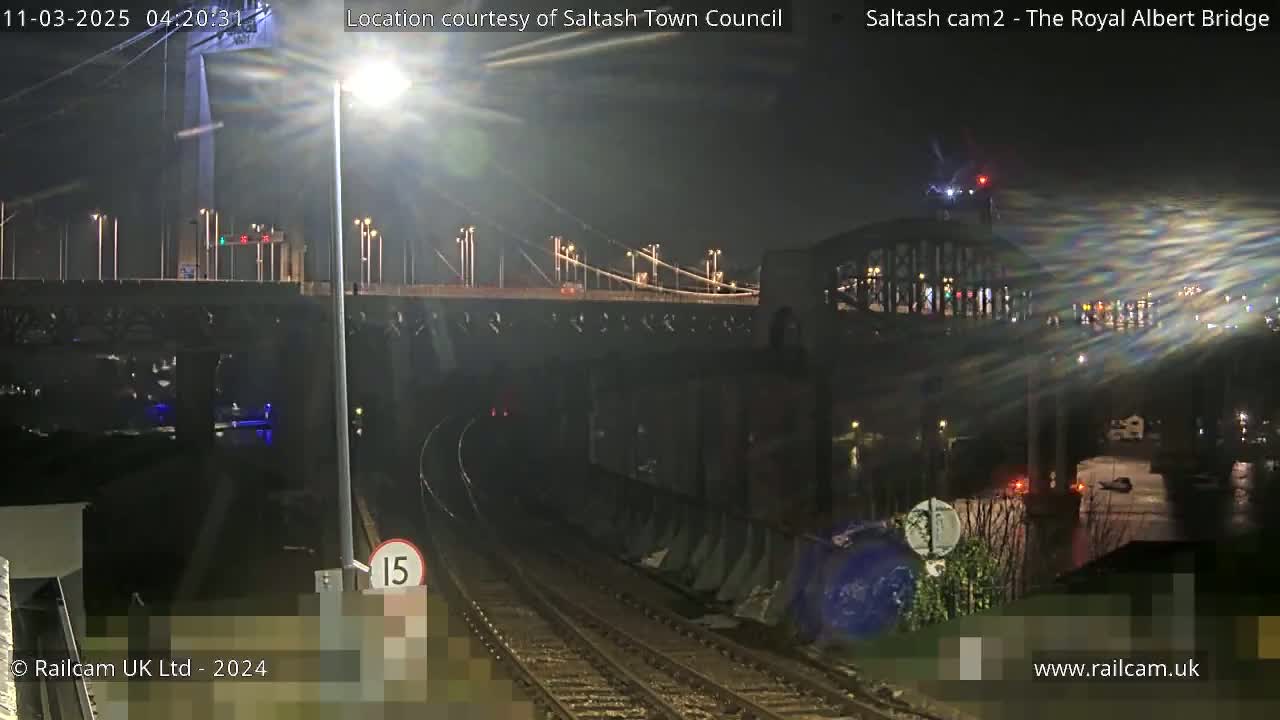 A nighttime view of a train track leading toward a bridge and tunnel under a larger bridge, illuminated by streetlights in wet conditions.