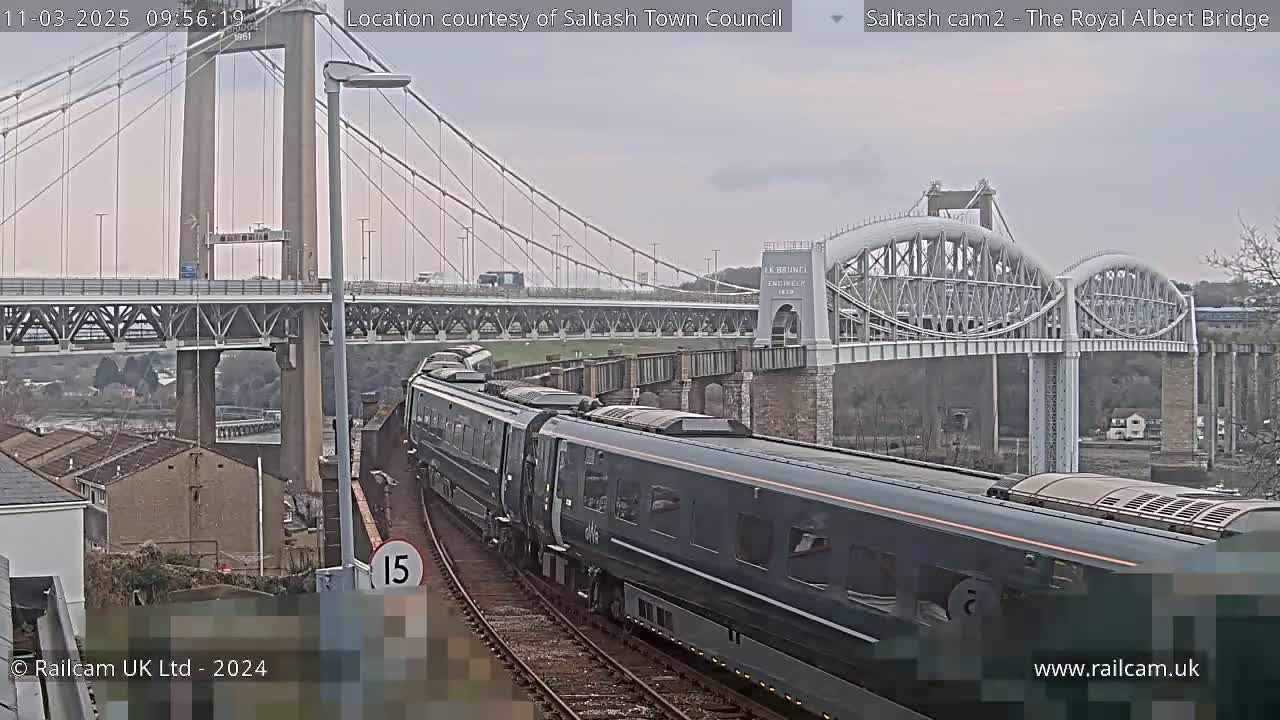 A dark-colored passenger train moves along train tracks below two bridges on an overcast day.