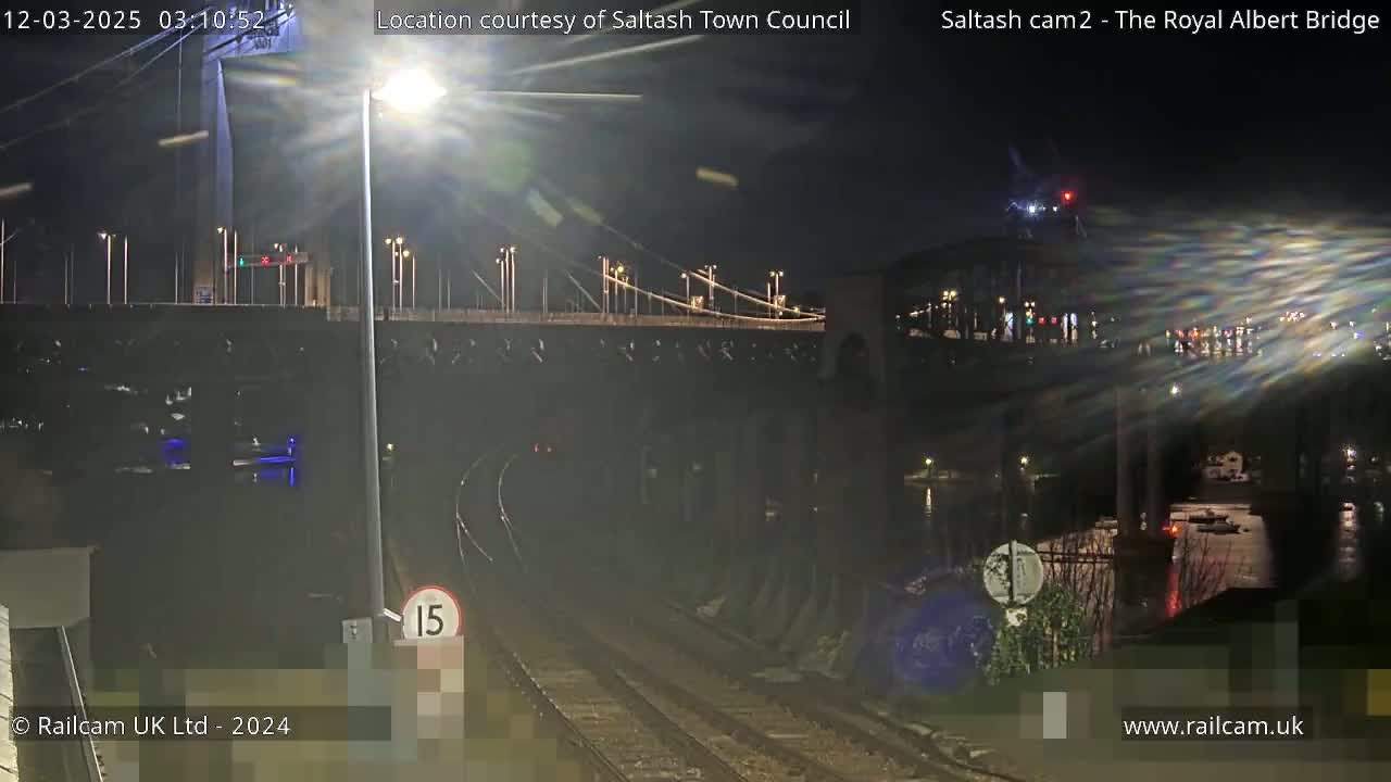 A nighttime view of a bridge over a body of water, with train tracks and a speed limit sign in the foreground.