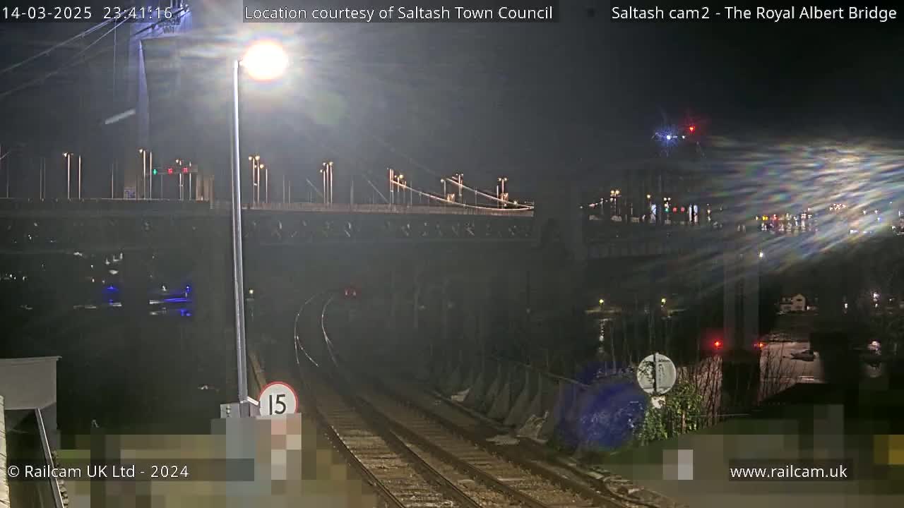 A nighttime view of train tracks leading toward a tunnel beneath a bridge, with lights illuminating the scene.