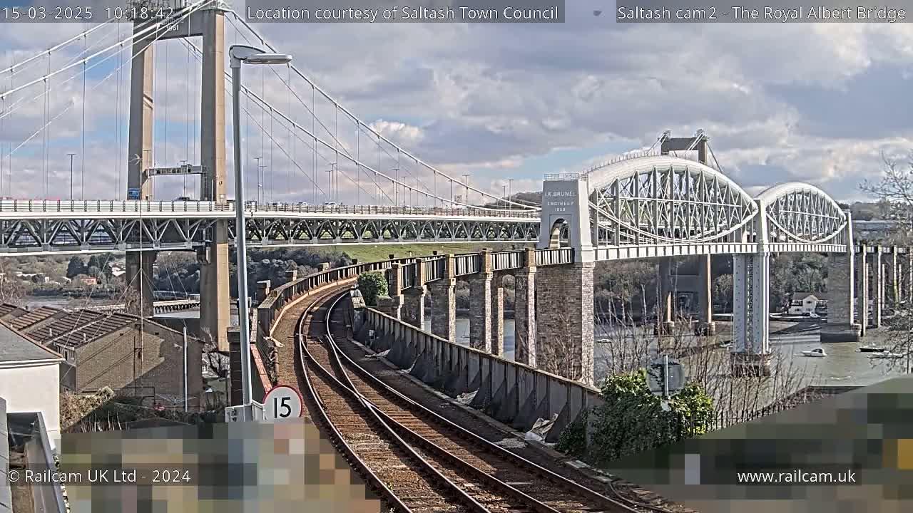A view of train tracks beside a river with two bridges, one a suspension bridge and the other an arch bridge, under a partly cloudy sky.