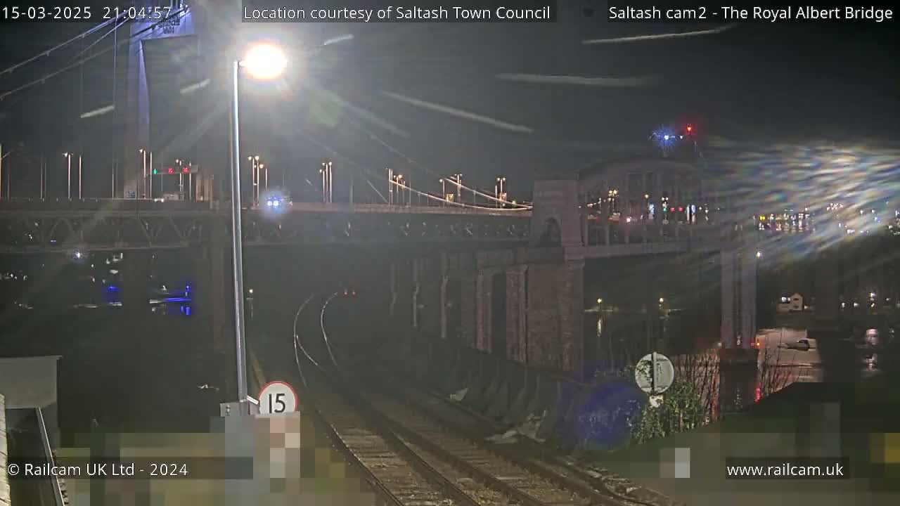 A nighttime view of a bridge over railroad tracks, with a streetlight illuminating the scene under partly cloudy skies.