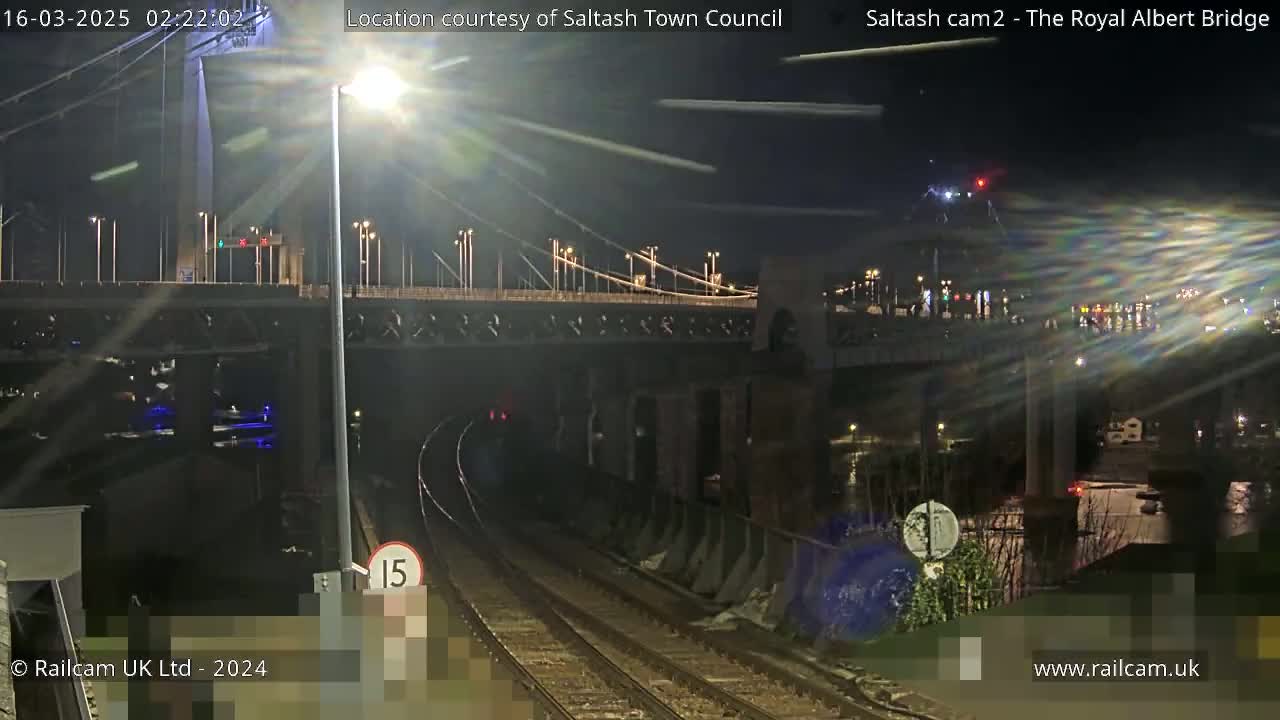 A nighttime view of a suspension bridge over train tracks, illuminated by streetlights under a clear, dark sky.