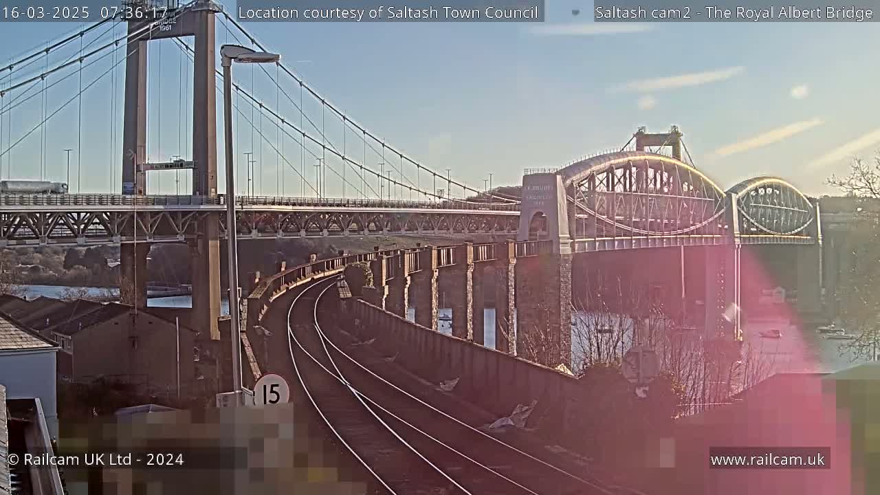 Two bridges span a river under a clear, sunny sky, with train tracks visible in the foreground.