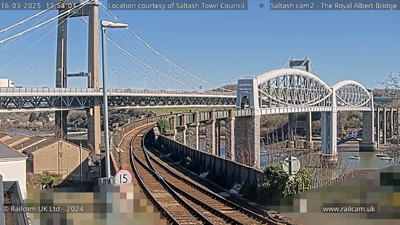 Under a clear blue sky, a railway line curves beneath two bridges spanning a waterway; one is a modern cable-stayed bridge, and the other is a historic arch bridge.
