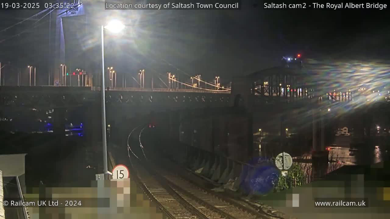A nighttime view of train tracks leading toward a bridge, illuminated by streetlights under a dark sky.