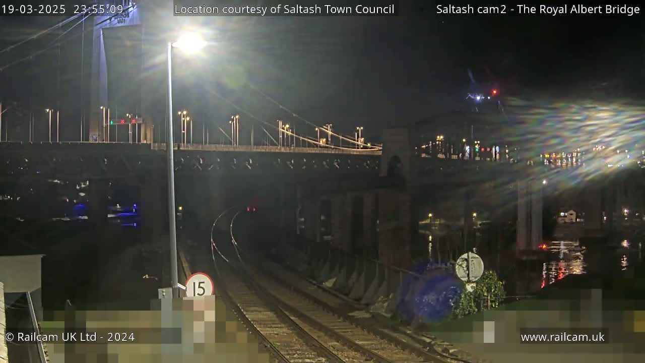 A nighttime view of a suspension bridge over water, with train tracks and a speed limit sign in the foreground.
