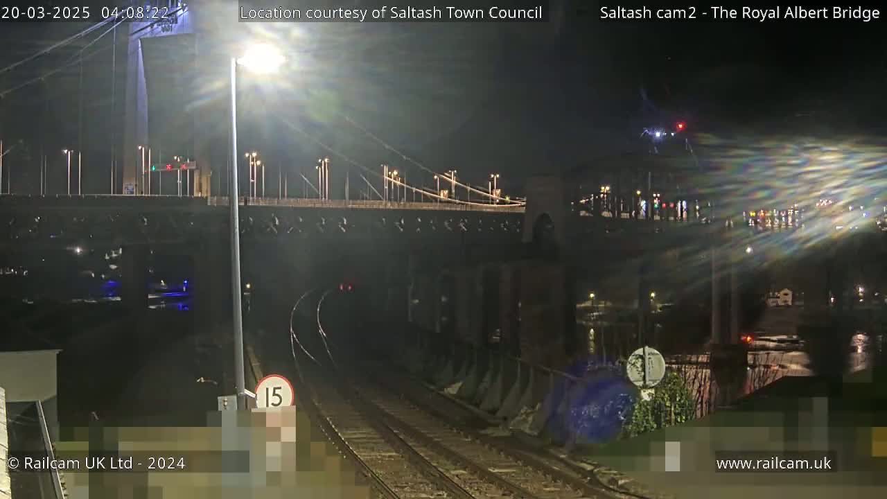 A nighttime view of a suspension bridge over a body of water, with train tracks and a speed limit sign in the foreground.