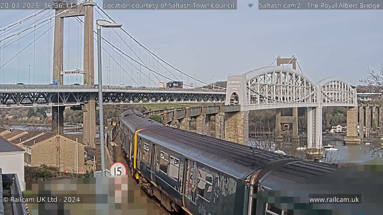 A train passes below a suspension bridge and a nearby arch bridge on a partly sunny day.