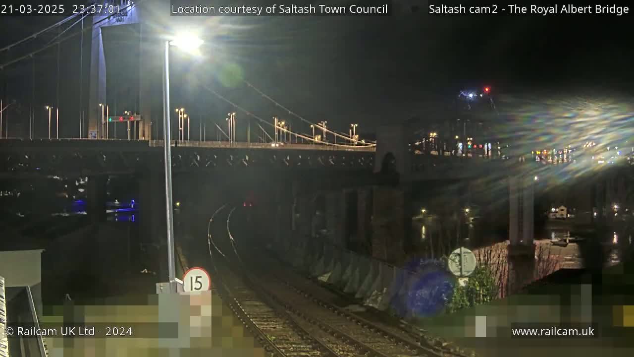 A nighttime view of a suspension bridge over a body of water, with train tracks and a speed limit sign visible in the foreground.