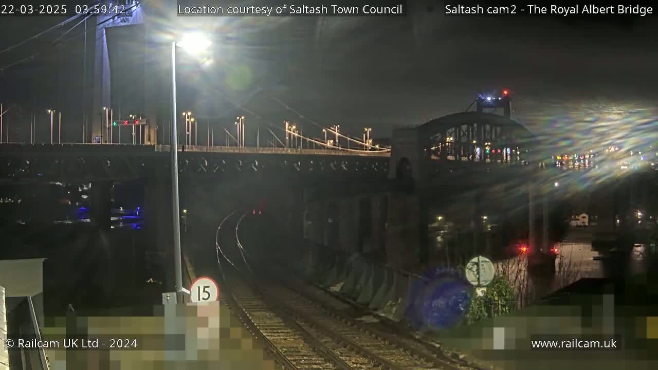 A nighttime view of a bridge over water with train tracks in the foreground under mostly dark skies.
