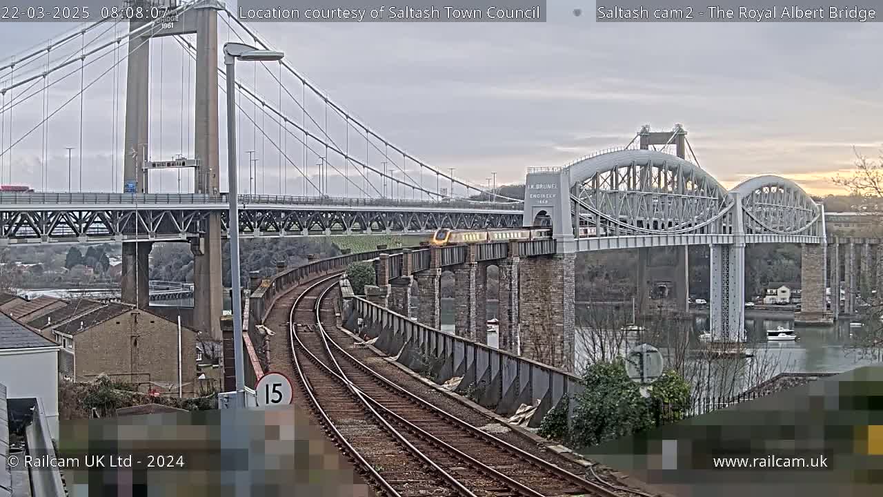 A train crosses a railway bridge next to a suspension bridge under an overcast sky.