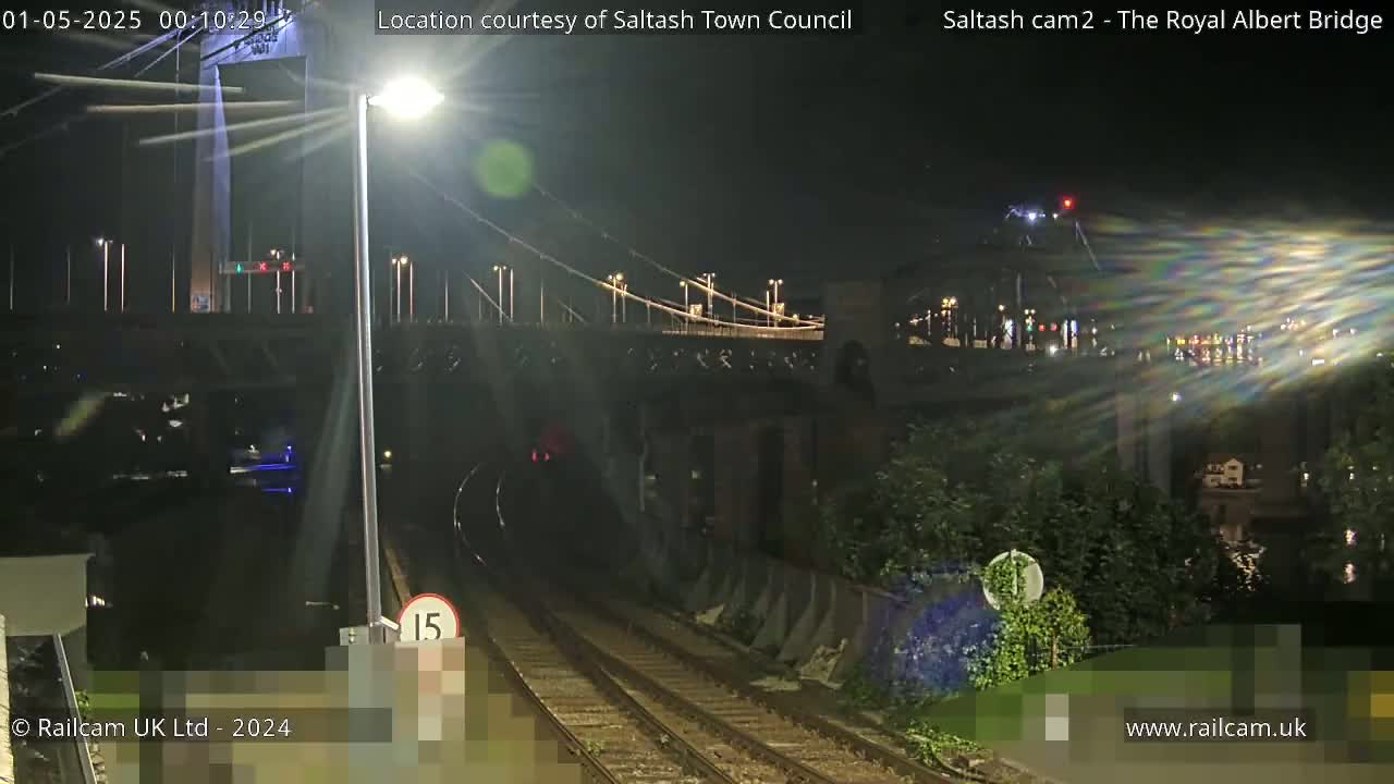 A nighttime view of a train approaching a tunnel beneath a suspension bridge, with lights illuminating the scene.
