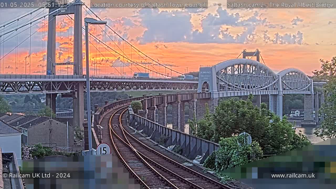 A sunset illuminates two bridges over a waterway, with train tracks visible in the foreground.