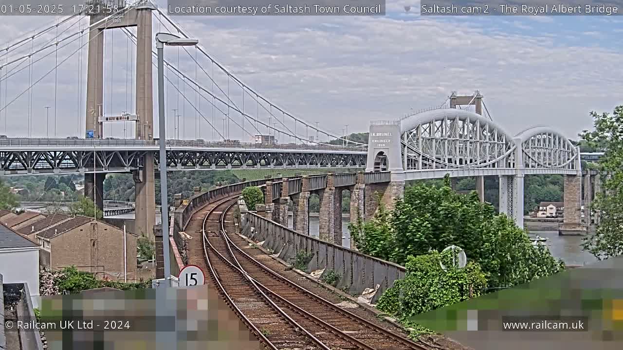 A view of train tracks curving toward two bridges, one a suspension bridge and the other an arch bridge, over a body of water under a cloudy sky.