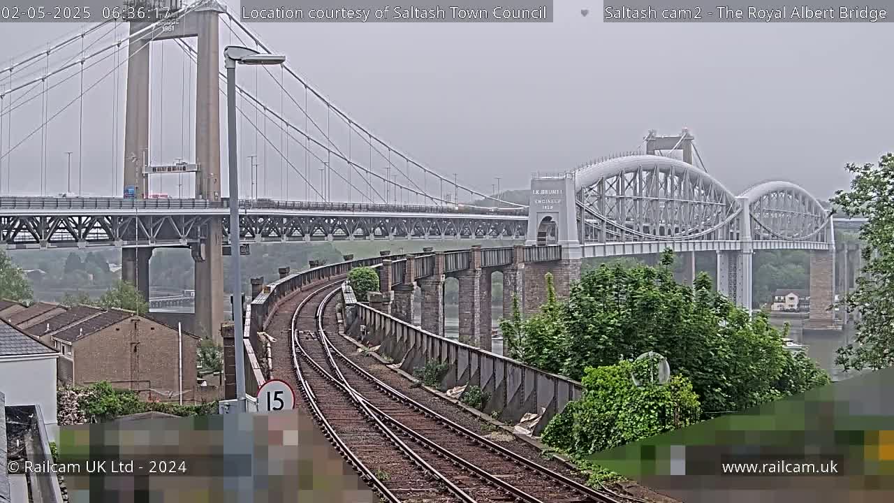 A view of train tracks leading to a railway bridge with another, larger suspension bridge visible in the background under an overcast sky.